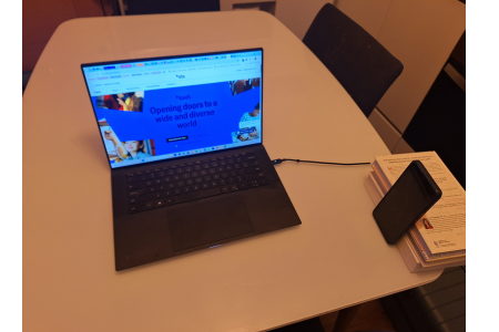 a laptop computer on a kitchen table with a cell phone propped by a stack of books