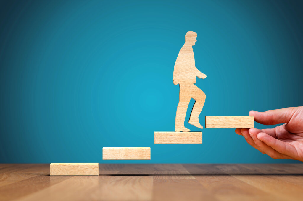 a wooden figure walking up a set of wooden blocks as steps with a hand holding out the next step