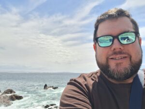 a selfie of a man with dark brown hair and sunglasses with the ocean in the background