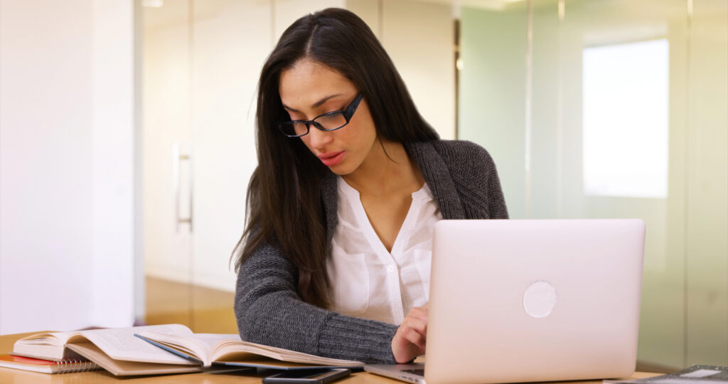 a woman is sitting at a desk looking down at a notebook and typing on her computer.