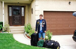 A teenager outside of a house wearing a suit jacket with some pins on it. At his feet are three big luggage pieces.