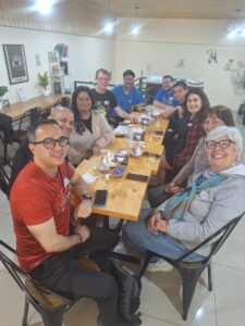 10 people seated around a long table in a coffee shop looking at the camera. They are men and women of different ages and nationalities, and there are some glasses of finished drinks on the table.