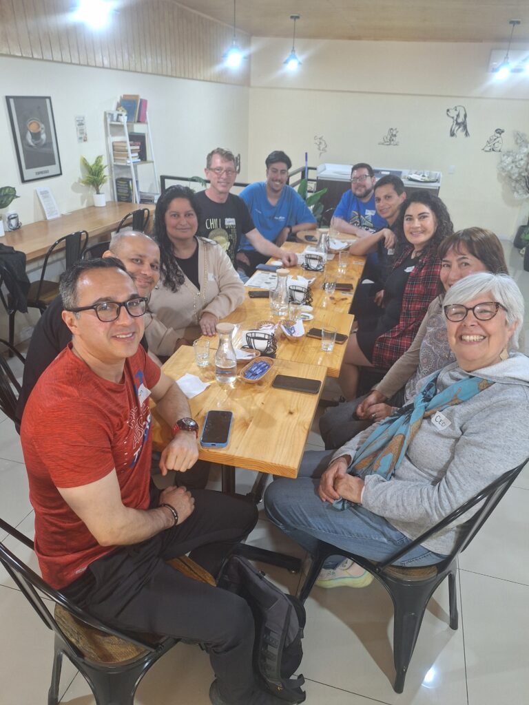10 people seated around a long table in a coffee shop looking at the camera. They are men and women of different ages and nationalities, and there are some glasses of finished drinks on the table.
